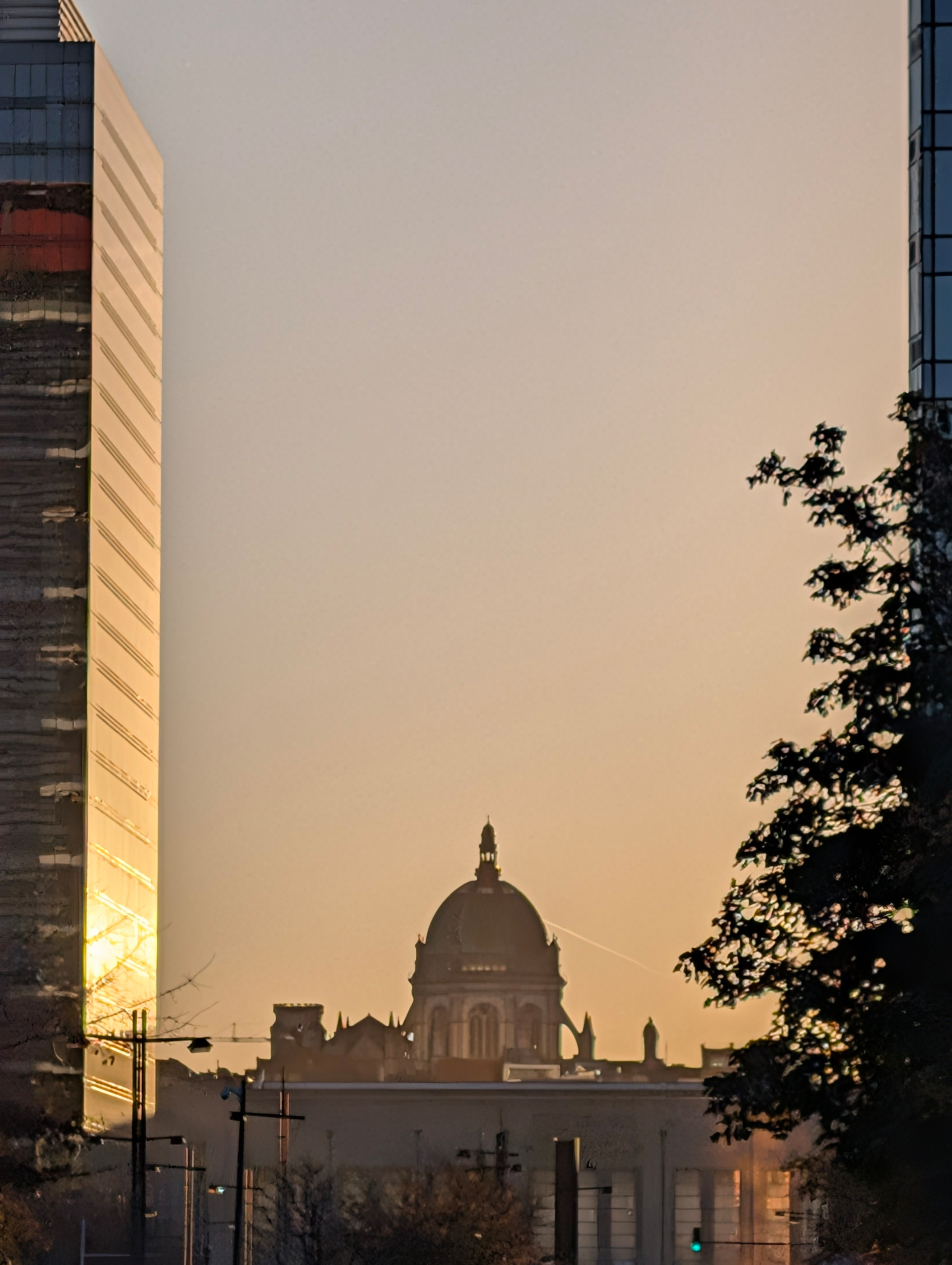 A domed building is framed by two tall structures and silhouetted trees under a warm, glowing sky.